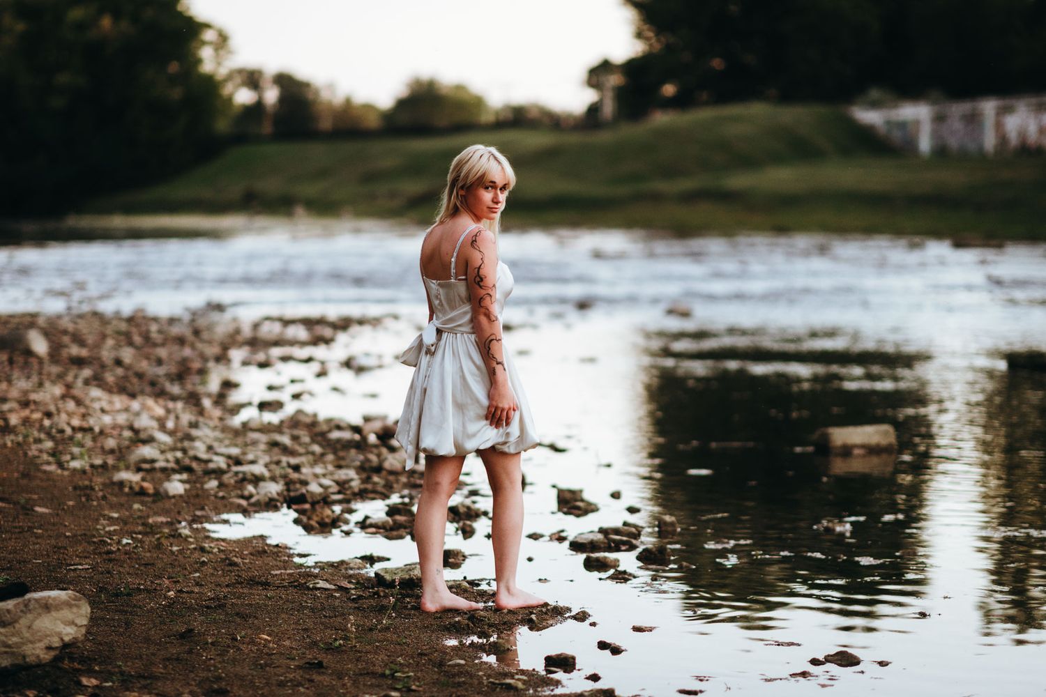 A person in a white dress stands by a serene lake shore during golden hour, creating a peaceful reflection in the water.