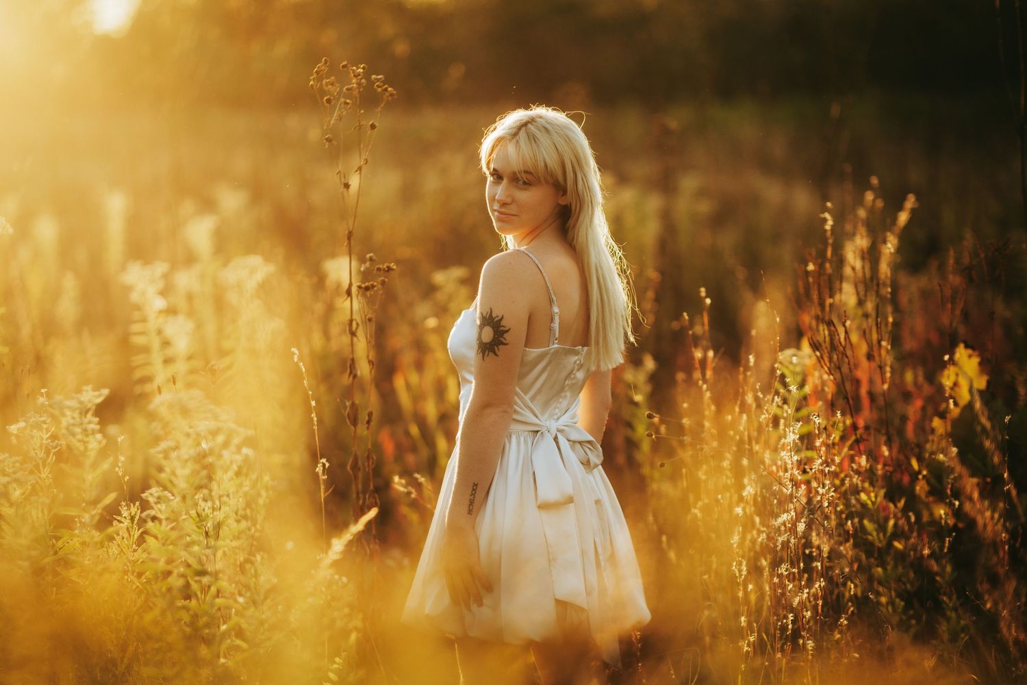 A figure in a white dress stands in a golden sunlit field of tall grass and wildflowers at sunset.