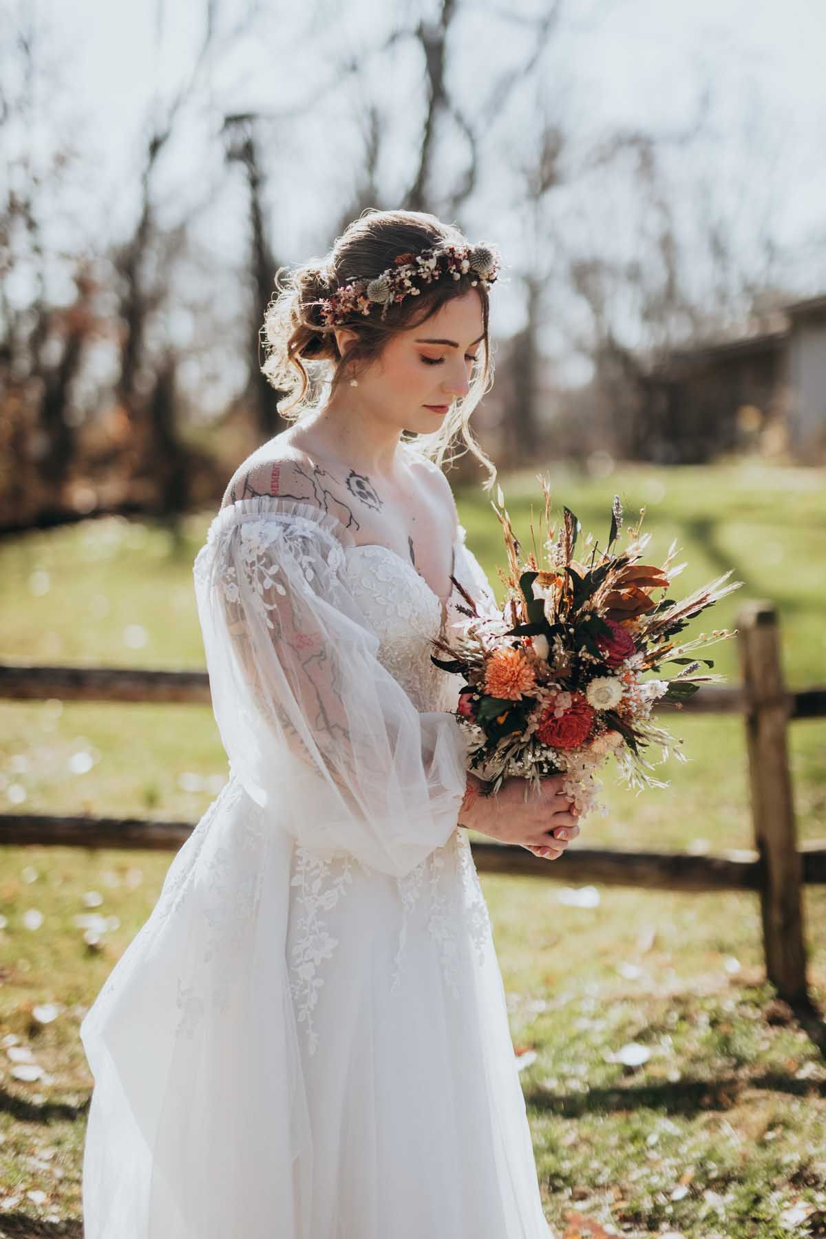 A bride in a lace-trimmed white gown holds an autumn bouquet in a sunlit outdoor setting with fallen leaves.