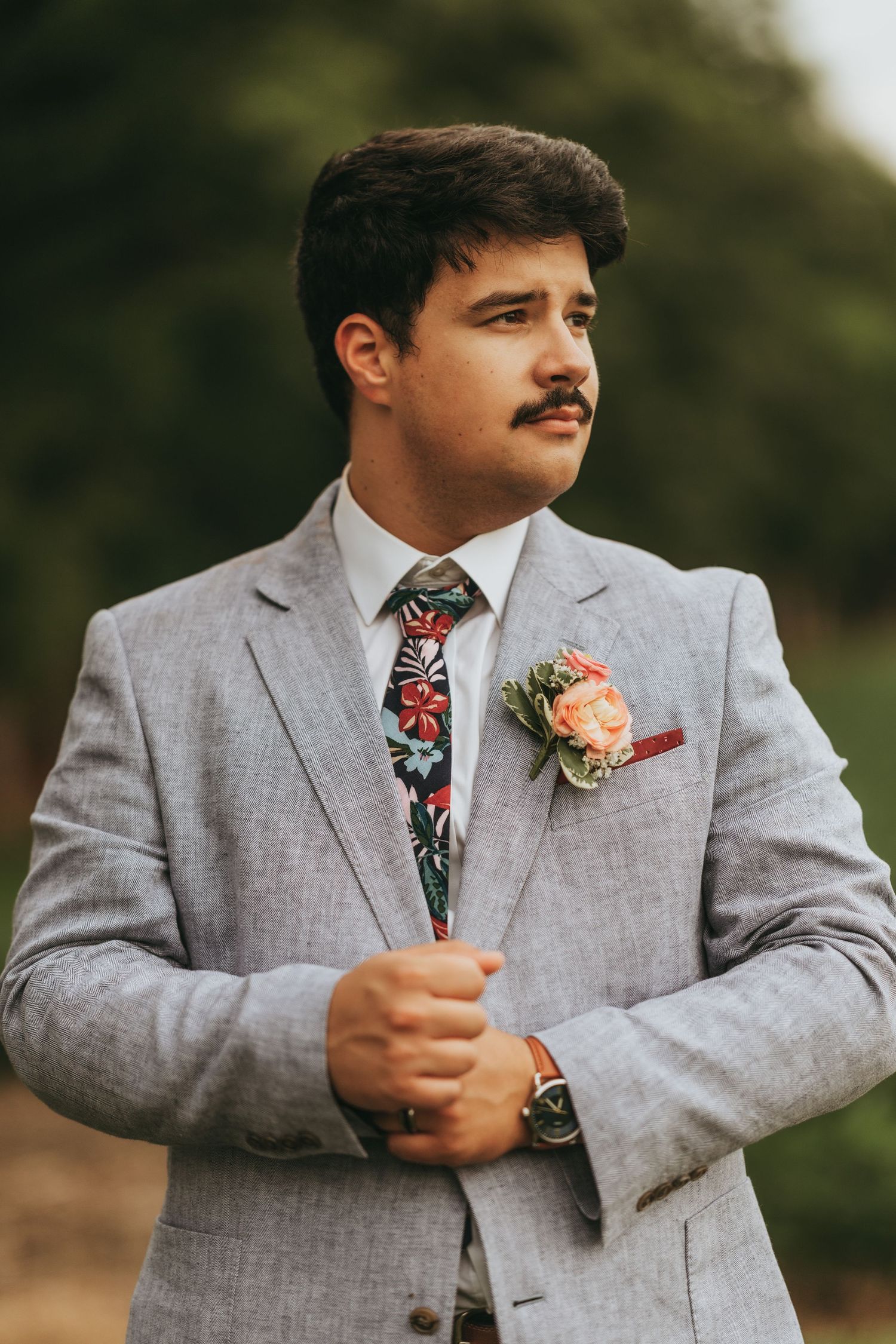 Handsome groom in gray suit with floral tie and boutonniere posing outdoors at wedding.