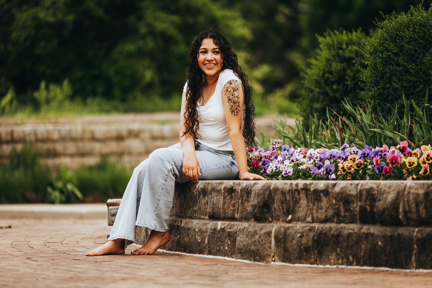 A person in white attire sits casually on stone steps surrounded by vibrant purple and pink flowers in a garden setting.