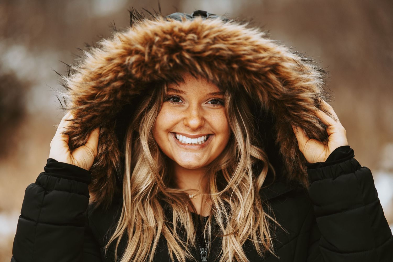 A person wearing a brown fur-trimmed hooded coat smiles warmly in a winter outdoor setting.