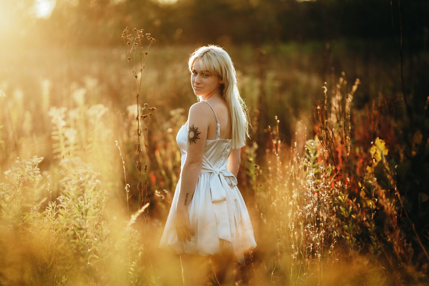 A person in a white dress stands in a golden sunlit meadow with tall grass and wildflowers at sunset.