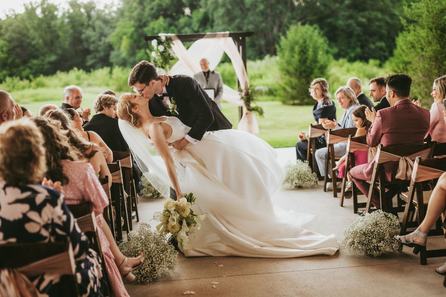 Bride and groom share a romantic kiss at outdoor wedding ceremony with guests seated along the aisle.