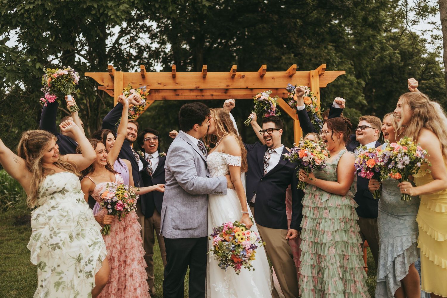 Wedding party celebrating with couple kissing under wooden pergola.
