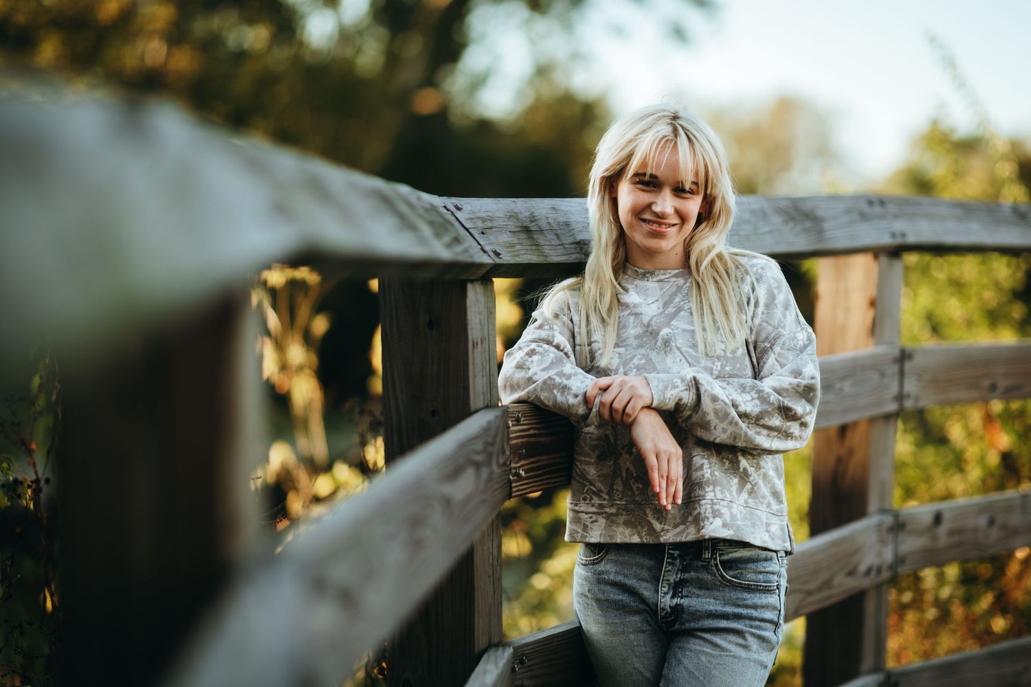 A person in a gray sweater sits on a wooden fence during golden hour with autumn foliage in the background.