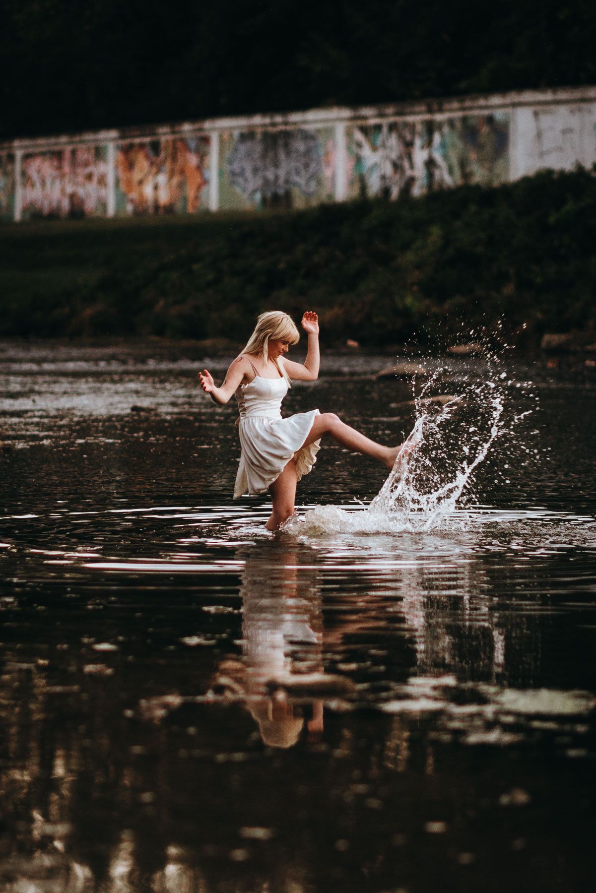 Person in white dress splashing and dancing in dark reflective water against graffiti wall backdrop at dusk.