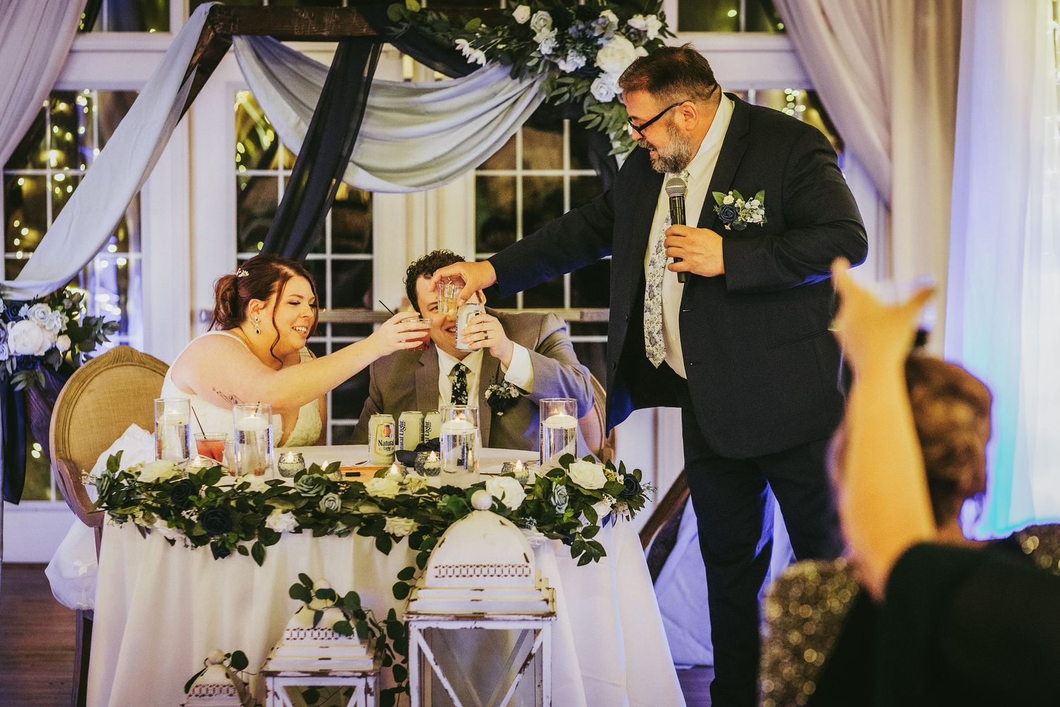 A wedding reception speech moment at a head table decorated with greenery and white linens.
