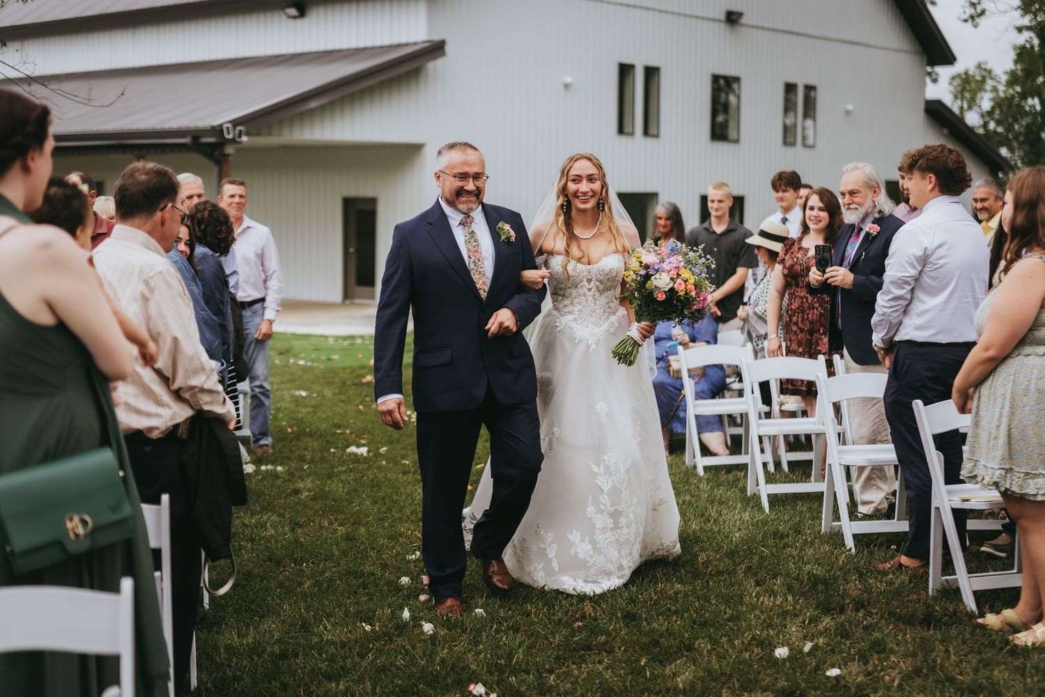 Bride walks down outdoor aisle with escort during sunny wedding ceremony at white barn venue.