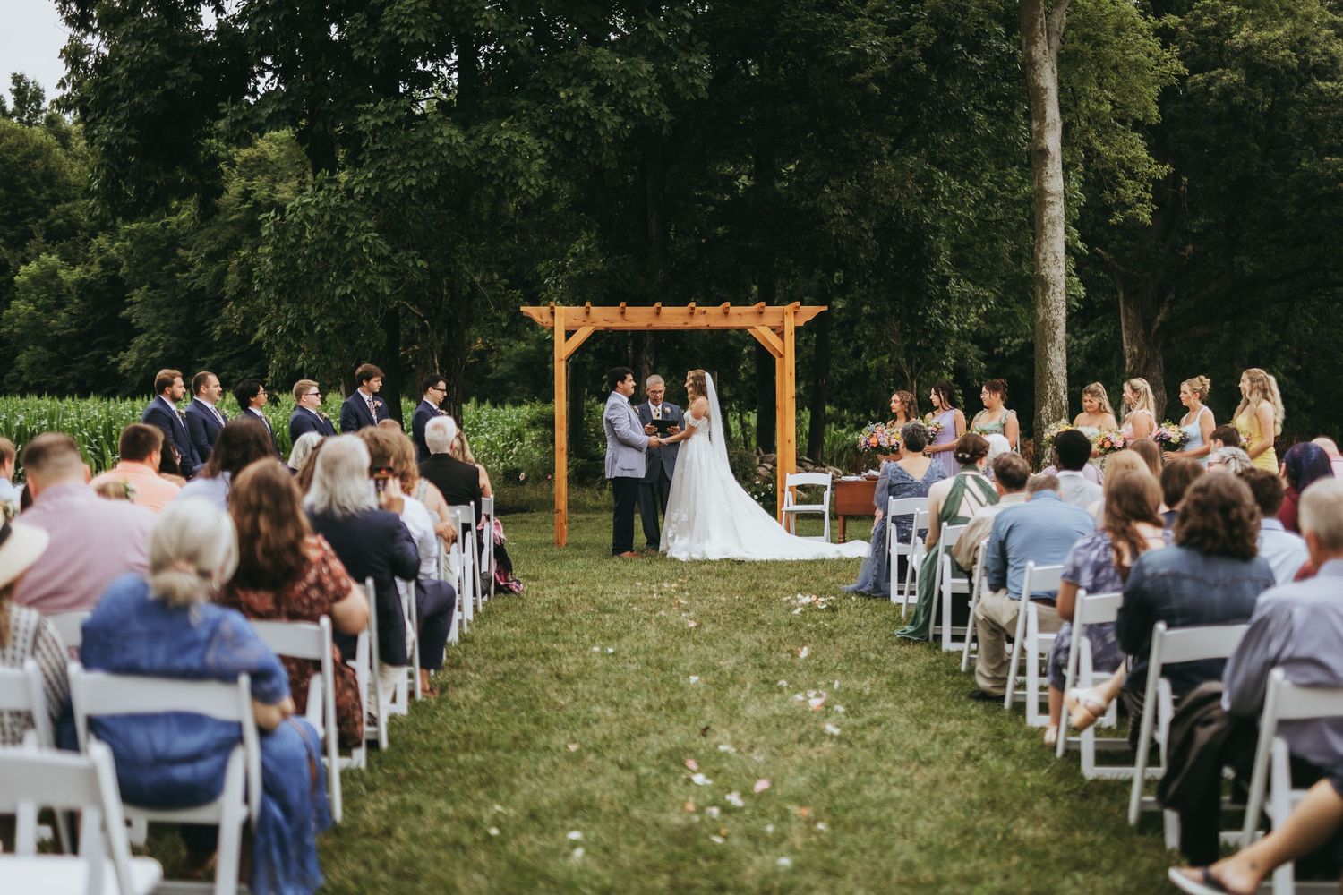 An outdoor wedding ceremony with guests seated in white chairs as a couple exchanges vows under a wooden pergola.