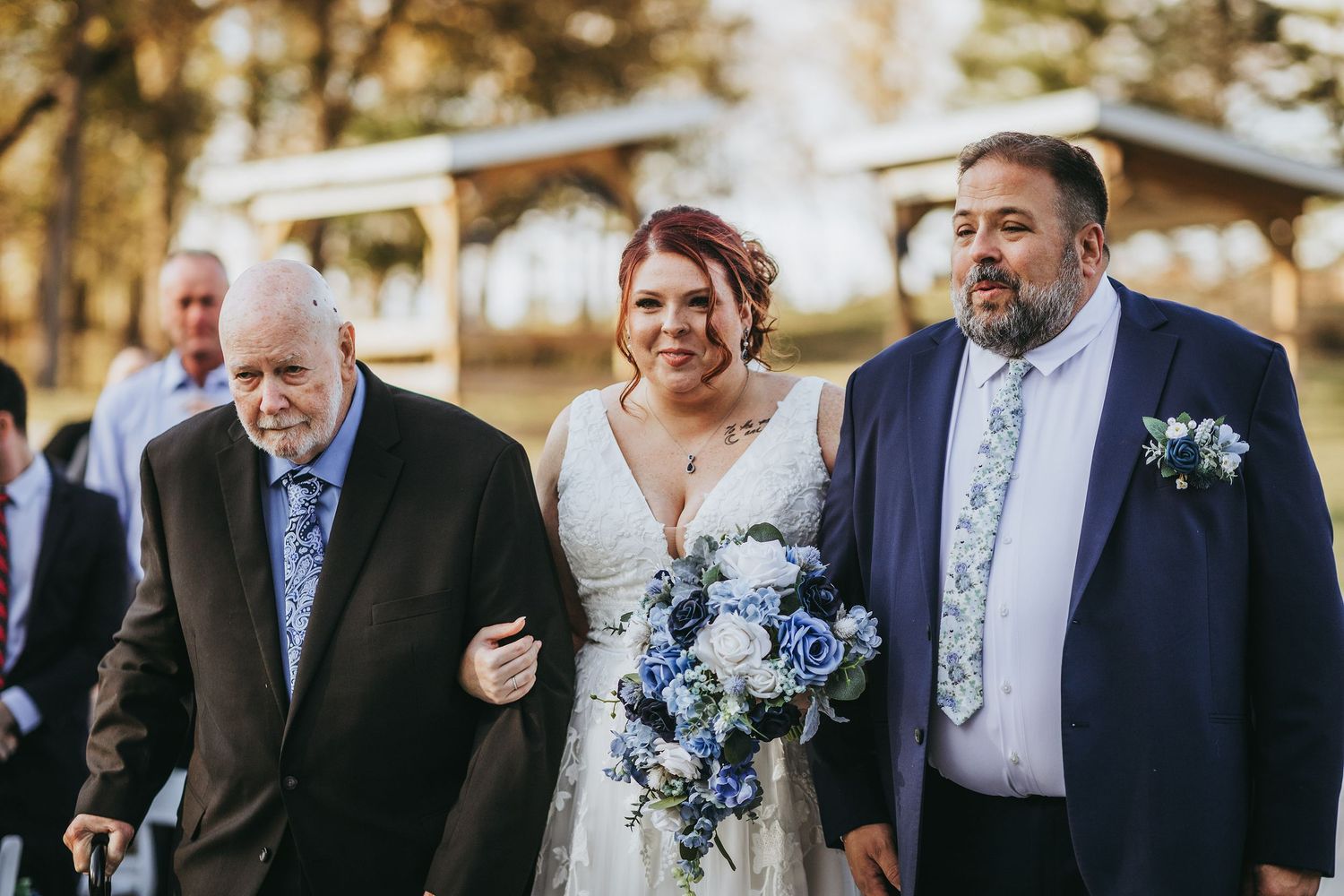 Wedding party members in navy and gray suits gather outdoors with blue floral bouquets.