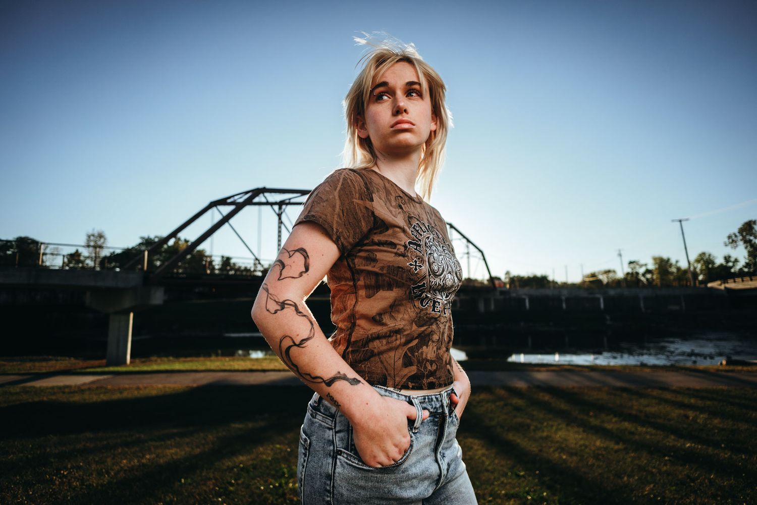 A person in a brown camo t-shirt and jeans poses outdoors against a sunset backdrop with a metal structure visible.