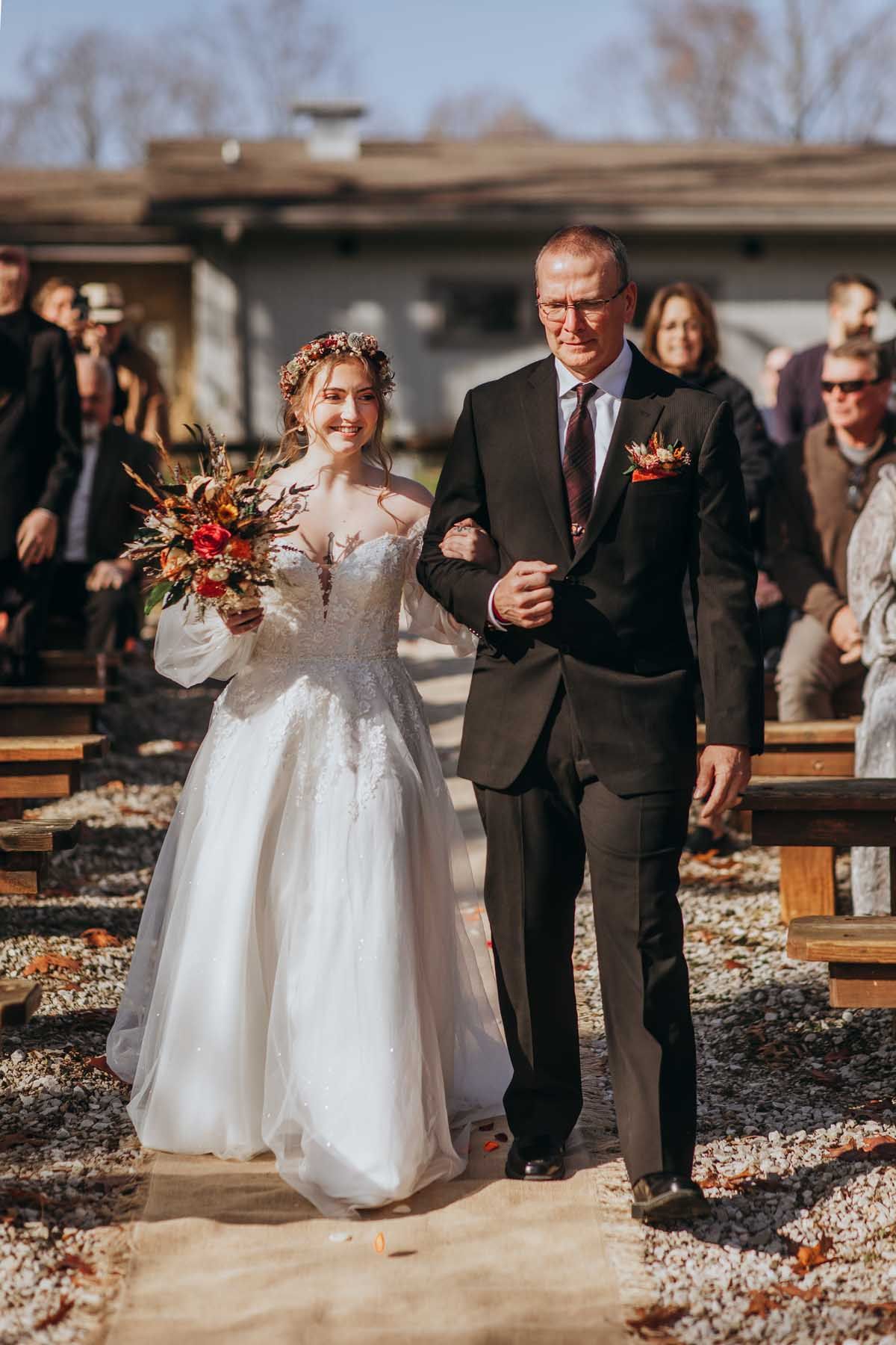 A wedding ceremony processional on a rustic outdoor path with fall foliage in the background.