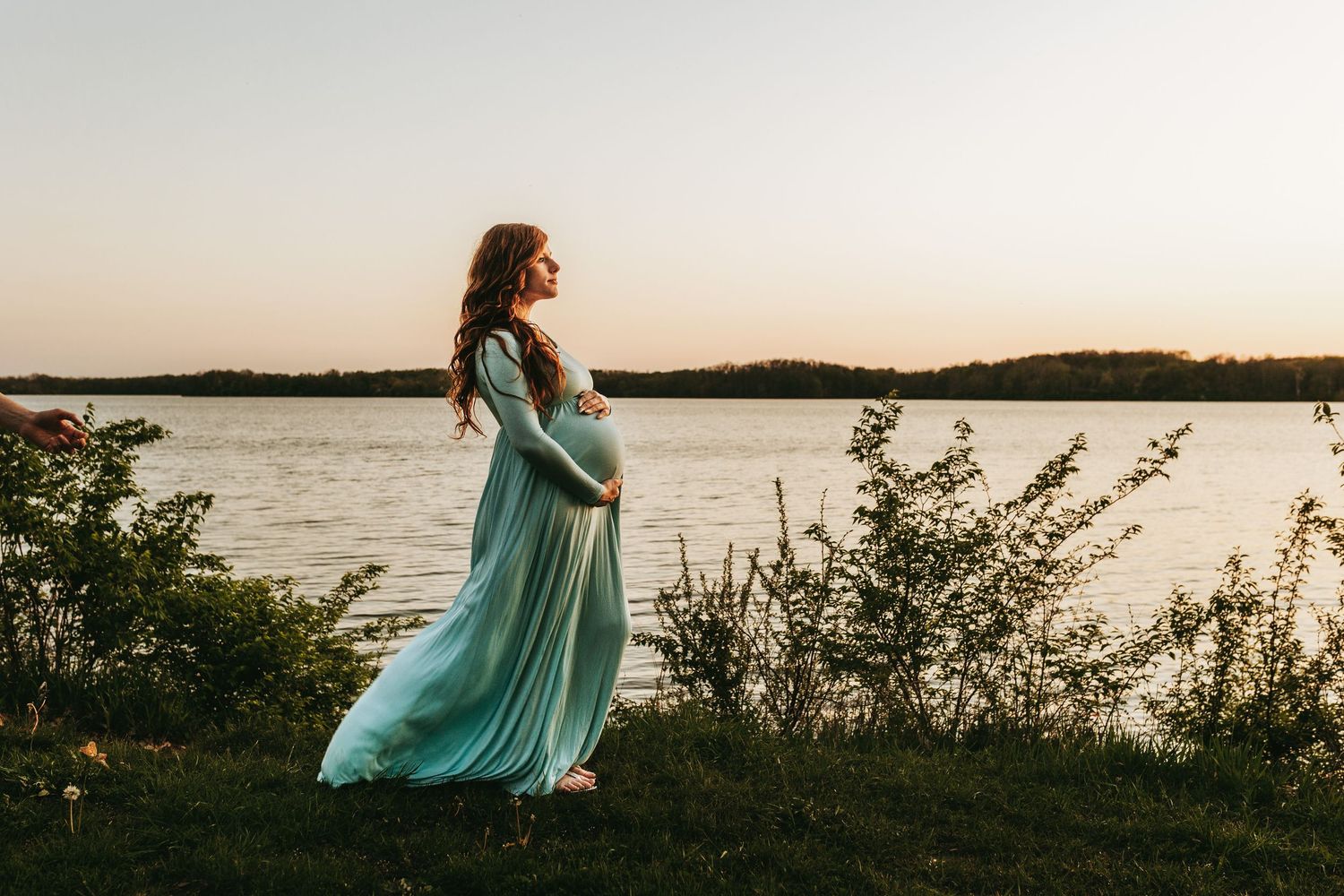 Silhouette of expectant mother in flowing turquoise dress standing by lakeside at sunset.