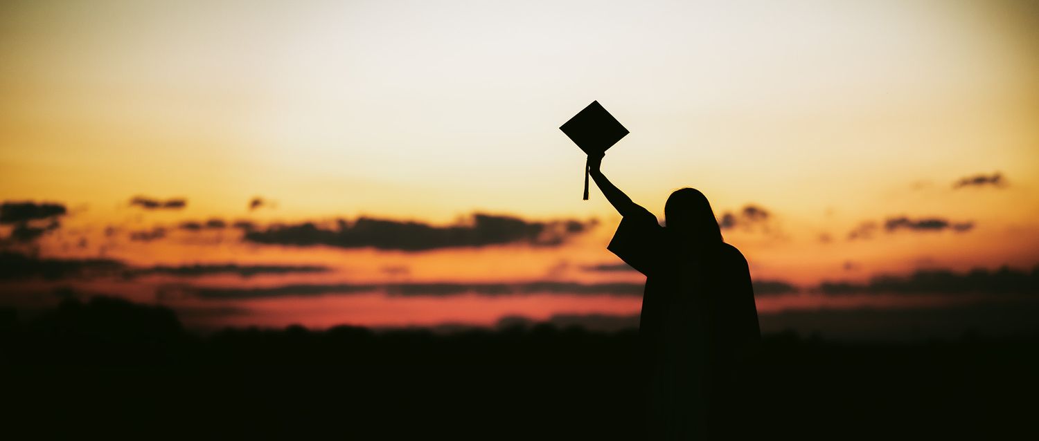 A silhouette raises their graduation cap against a vibrant sunset sky.