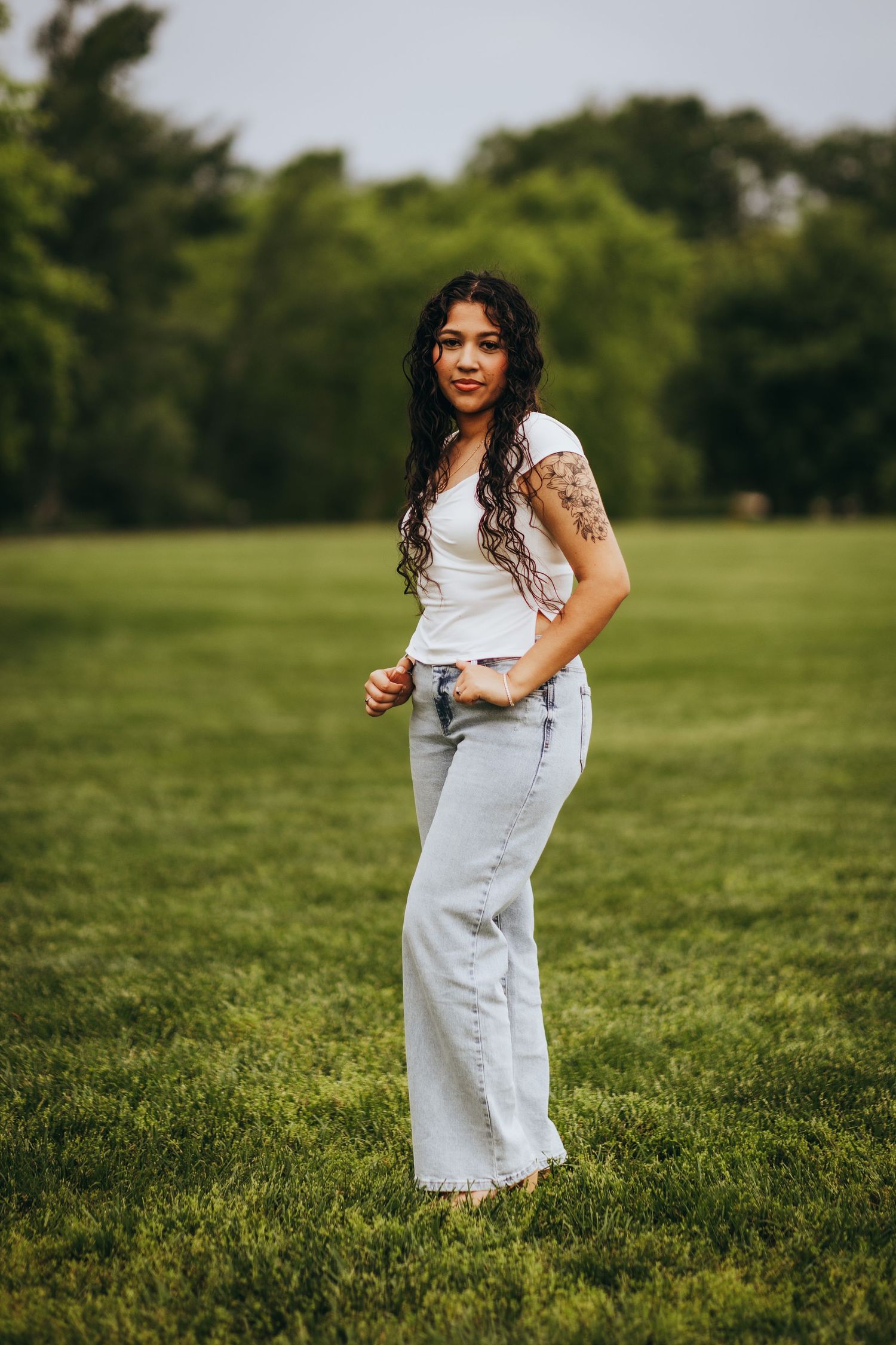 A person in a white top and light-colored overalls poses playfully in a sunny green field on a summer day.