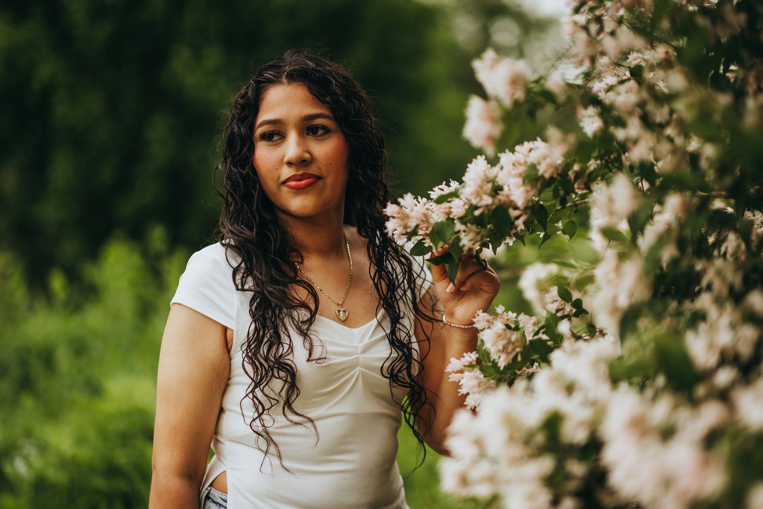 Woman in white dress poses gracefully among blooming cherry blossoms in a spring garden setting.