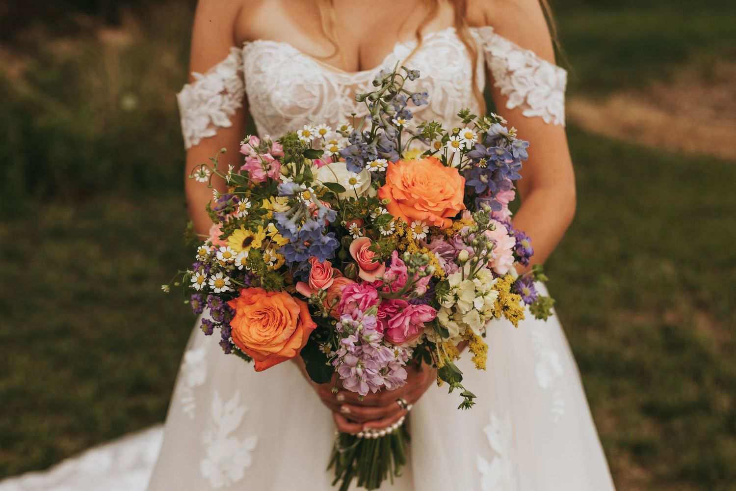 Bride holding vibrant wildflower bouquet with orange, pink, and purple blooms in lace wedding gown.
