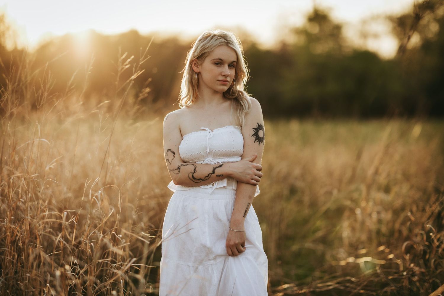 Blonde woman with tattoos in white dress standing in golden field at sunset.
