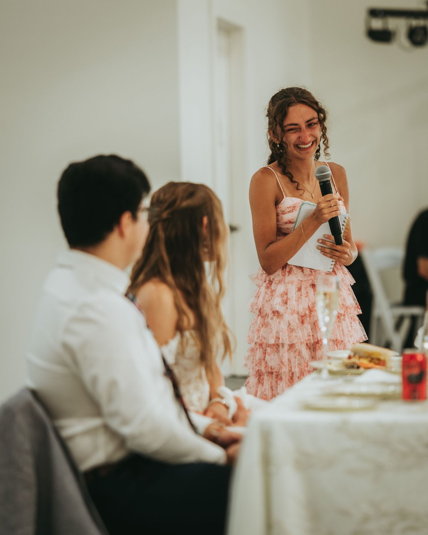 Joyful bridesmaid in pink ruffled skirt giving speech with microphone at elegant wedding reception.