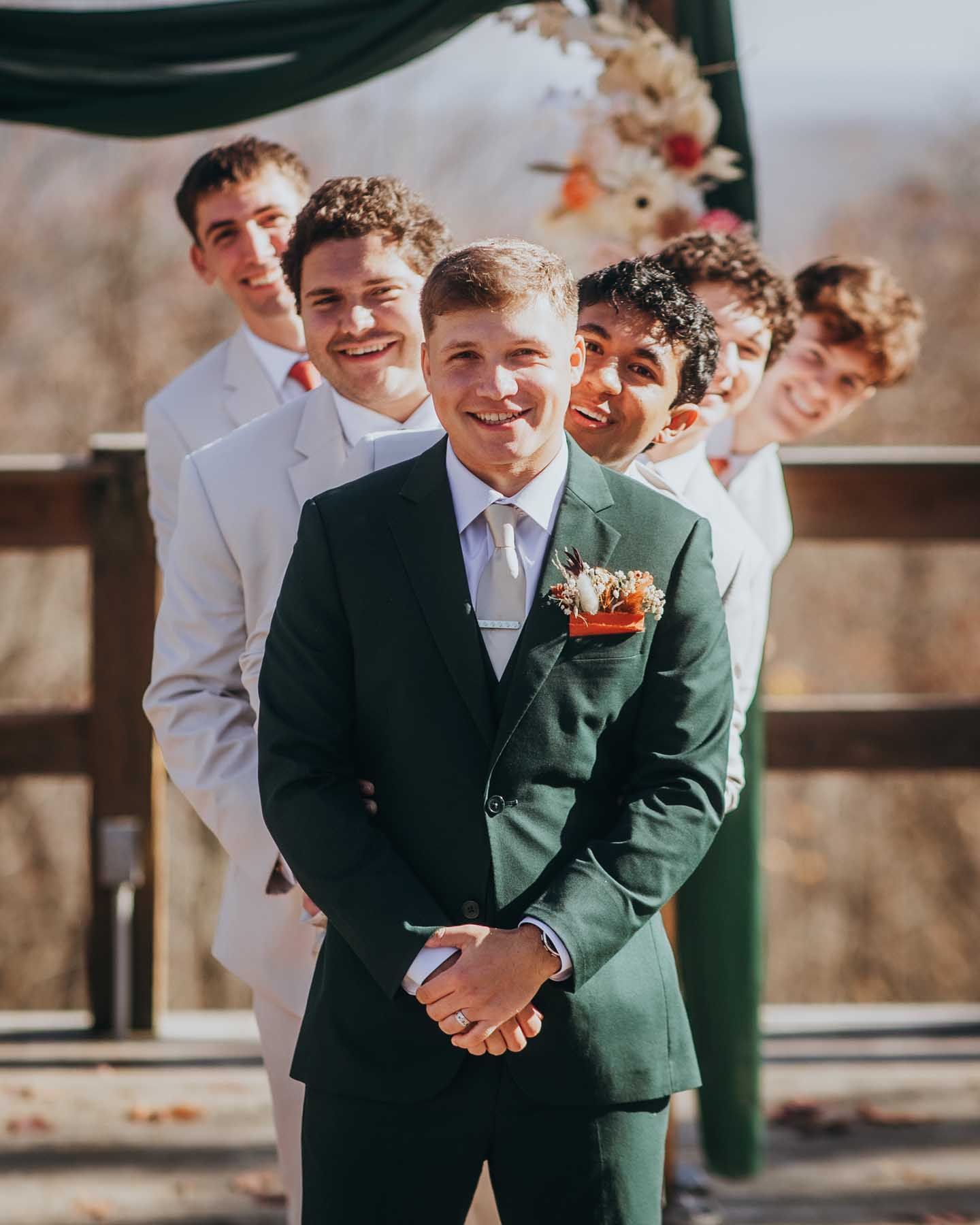 Groomsmen in dark suits and white ties stand in line outdoors on a sunny day, sharing candid laughter together.