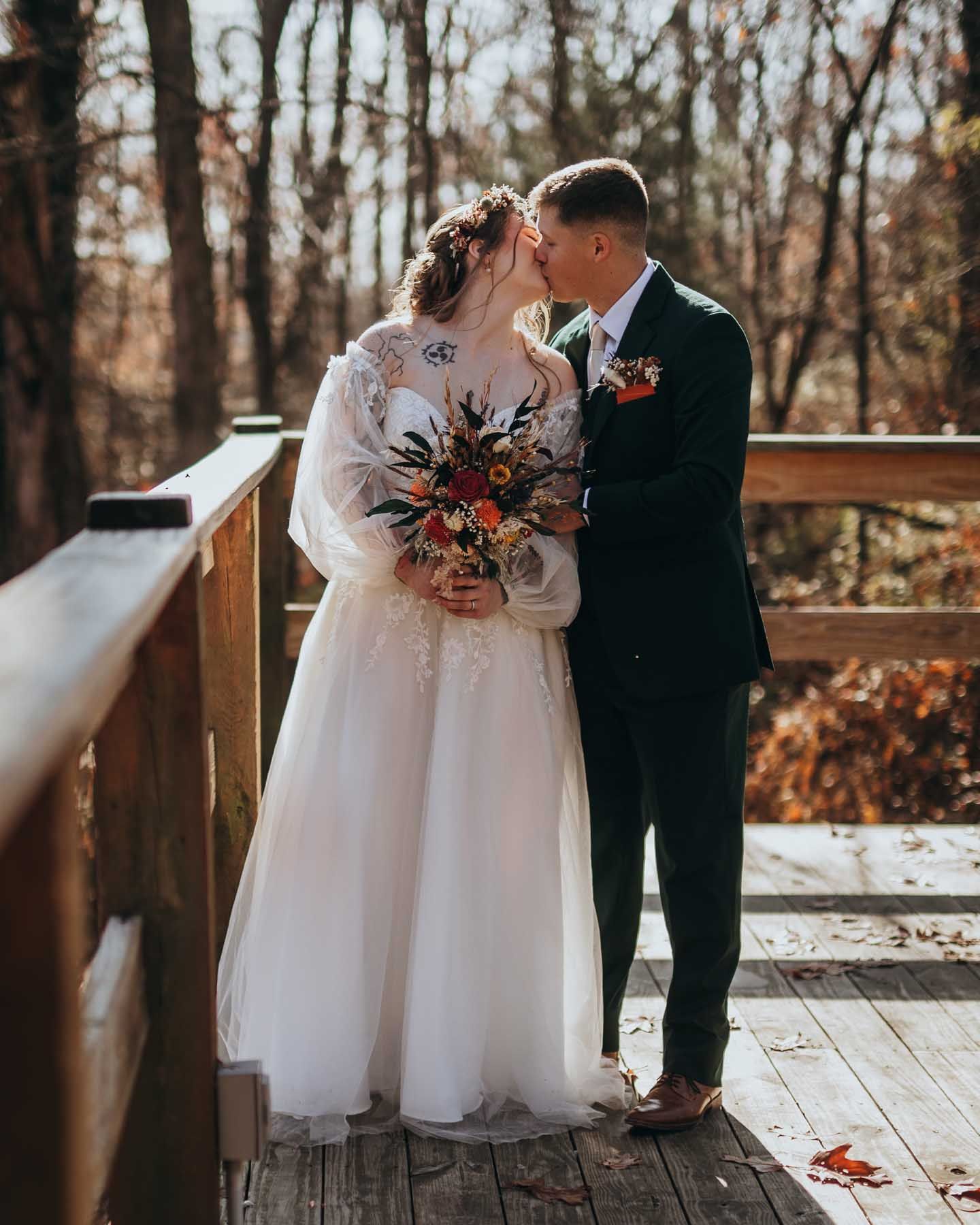 Wedding couple poses on a rustic wooden deck surrounded by autumn trees and scattered snow patches.