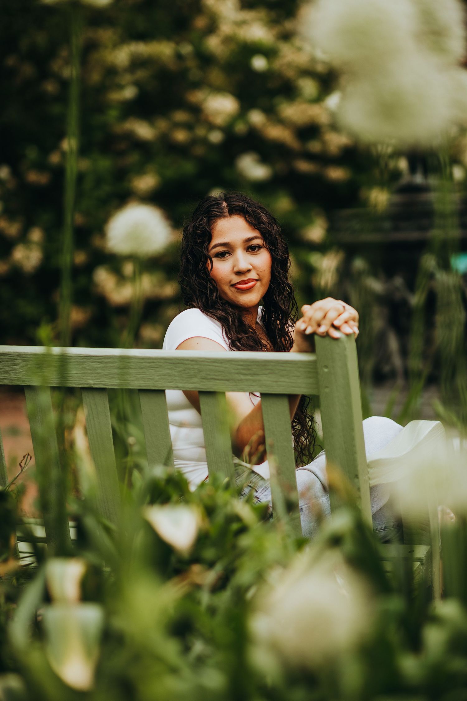 A person in a white shirt smiles while sitting behind a wooden fence surrounded by soft bokeh and greenery.