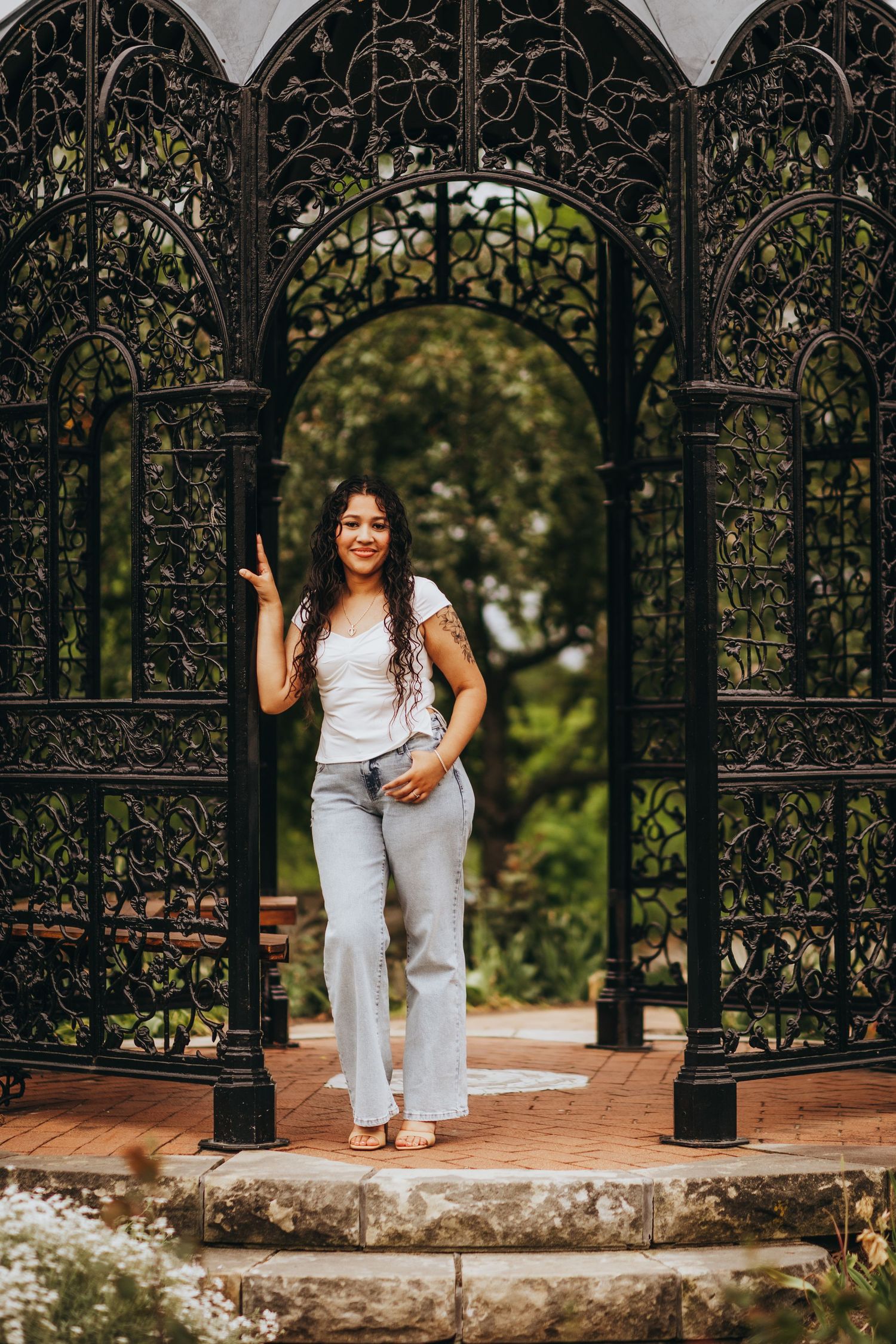 Two people in white outfits pose playfully by ornate black metal gates in a garden setting.