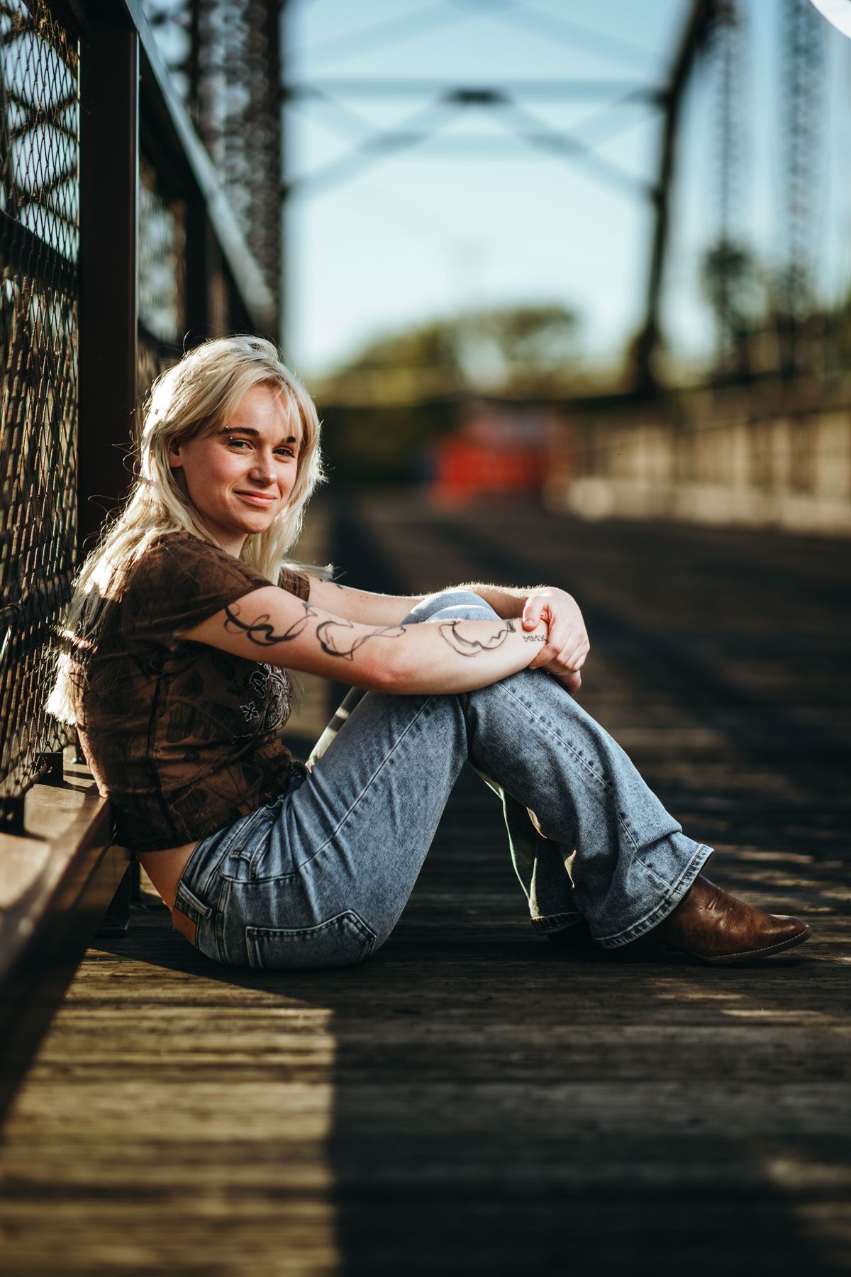 Casual portrait of someone sitting on wooden bridge wearing jeans and boots in warm sunset lighting.