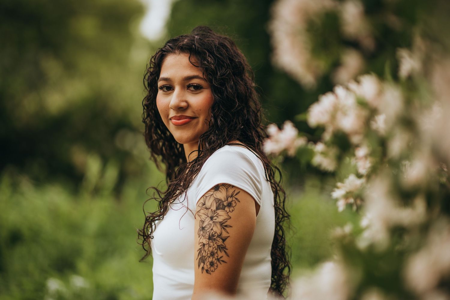 A woman with curly dark hair and a floral tattoo poses outdoors in a white dress against a blurred natural background.