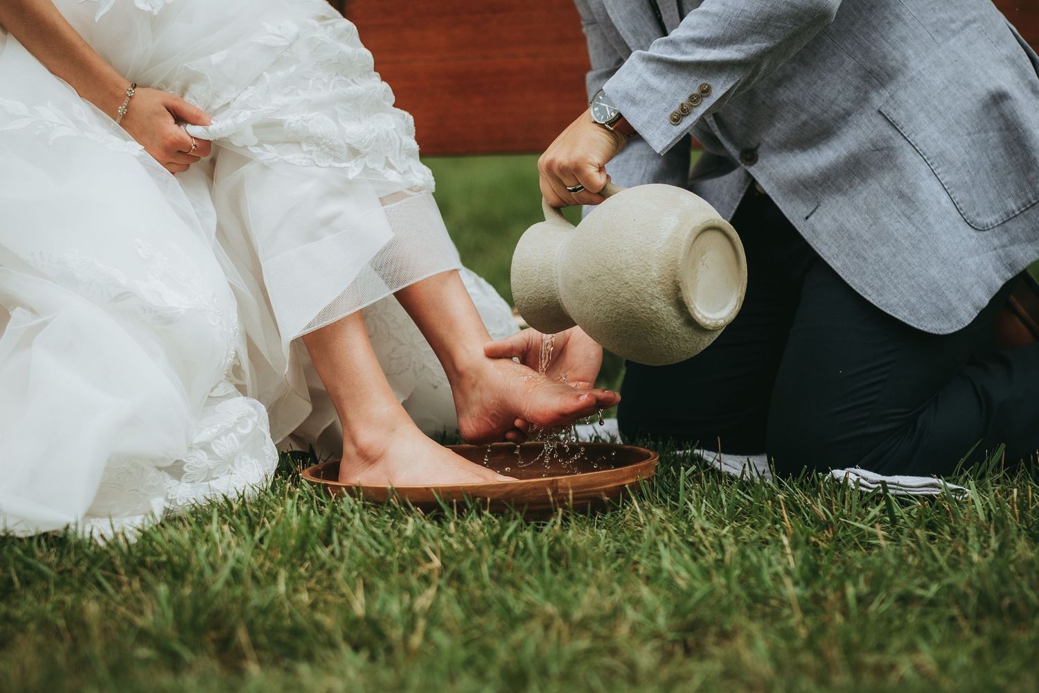 Groom performing foot washing ceremony on bride outdoors on grass.
