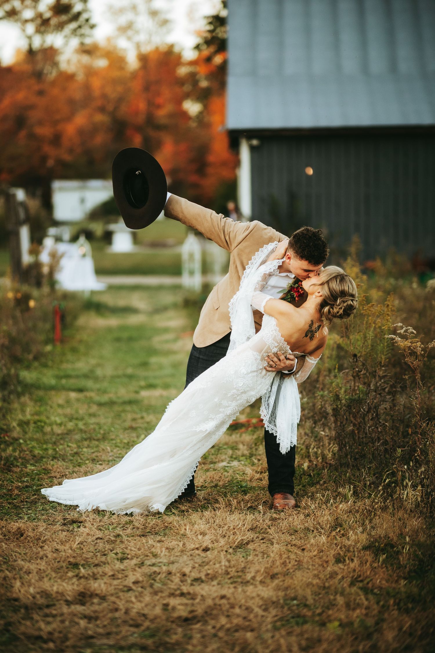 A romantic couple in wedding attire share a dramatic dip pose in front of a barn during autumn.