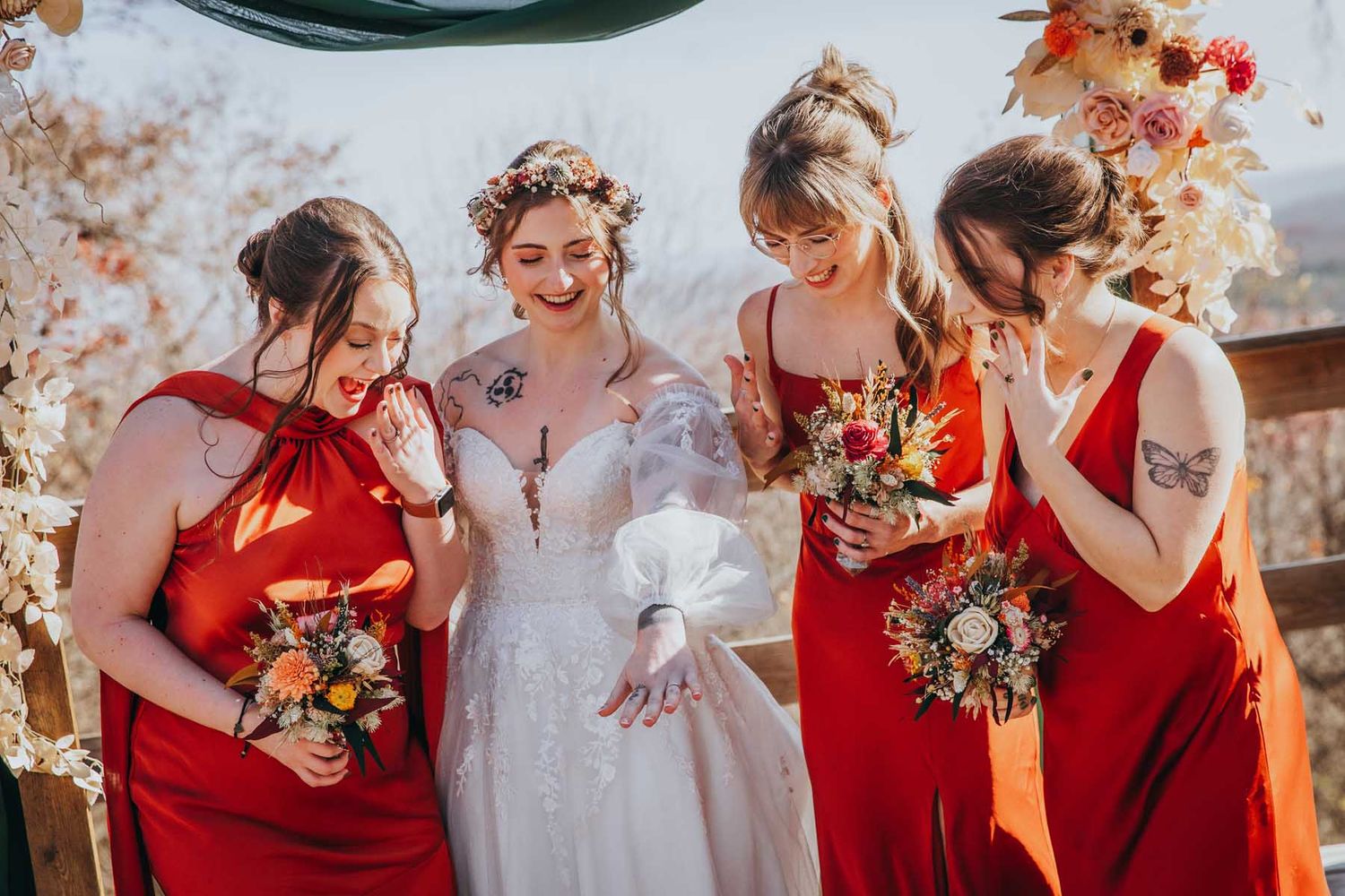 Bridesmaids in vibrant red dresses gather together outdoors, holding autumn-colored floral bouquets.