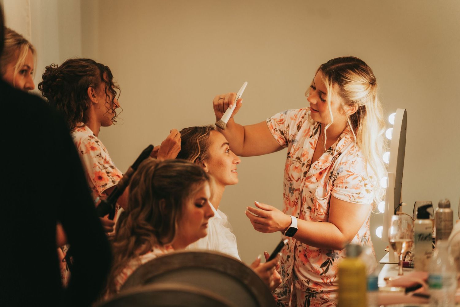 Bridesmaids getting hair and makeup done before wedding ceremony in warmly lit dressing room.