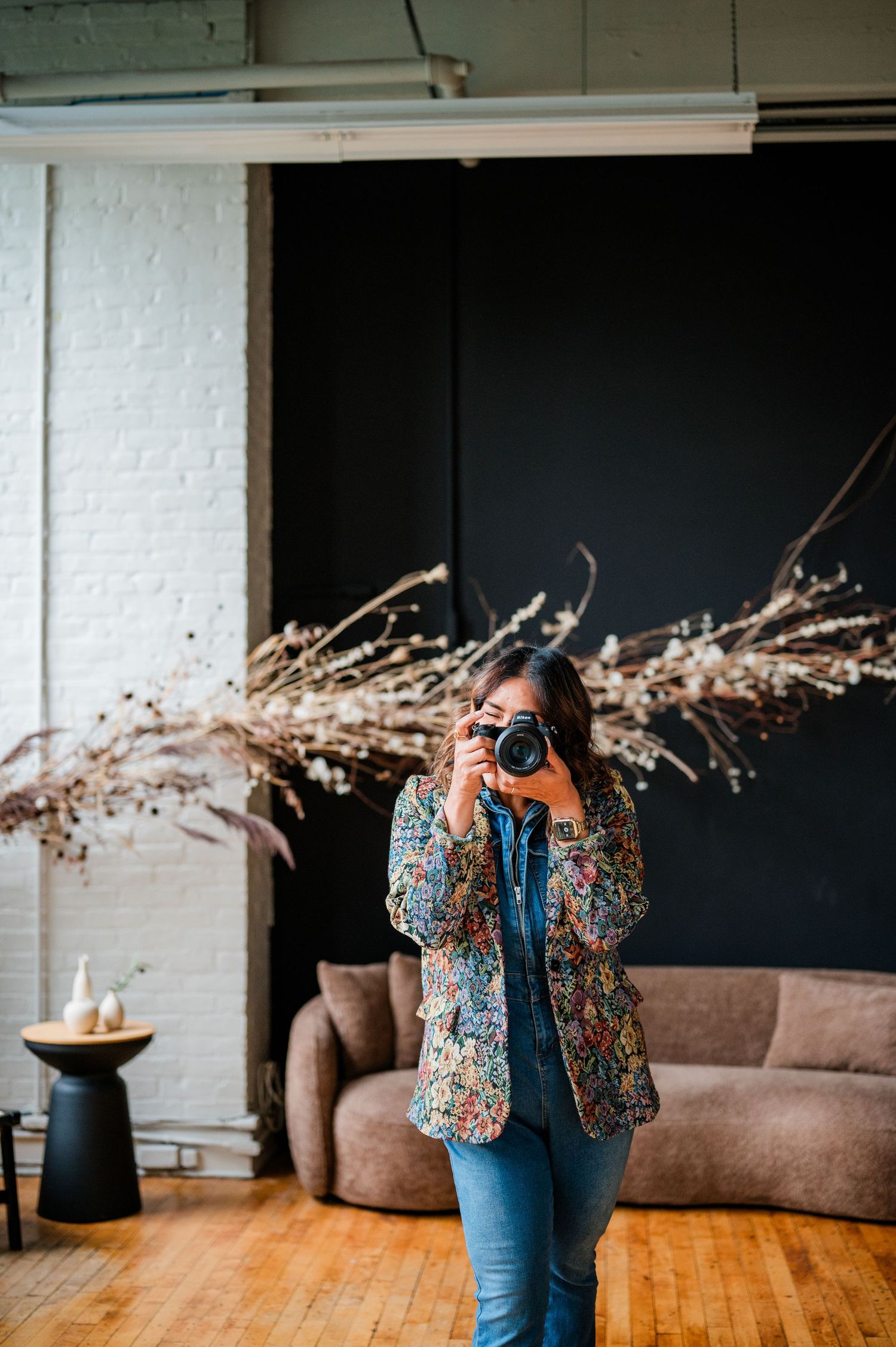 Woman in floral blazer photographing with a Nikon mirroless camera in modern studio with dried botanicals and dark accent wall Hartford, CT