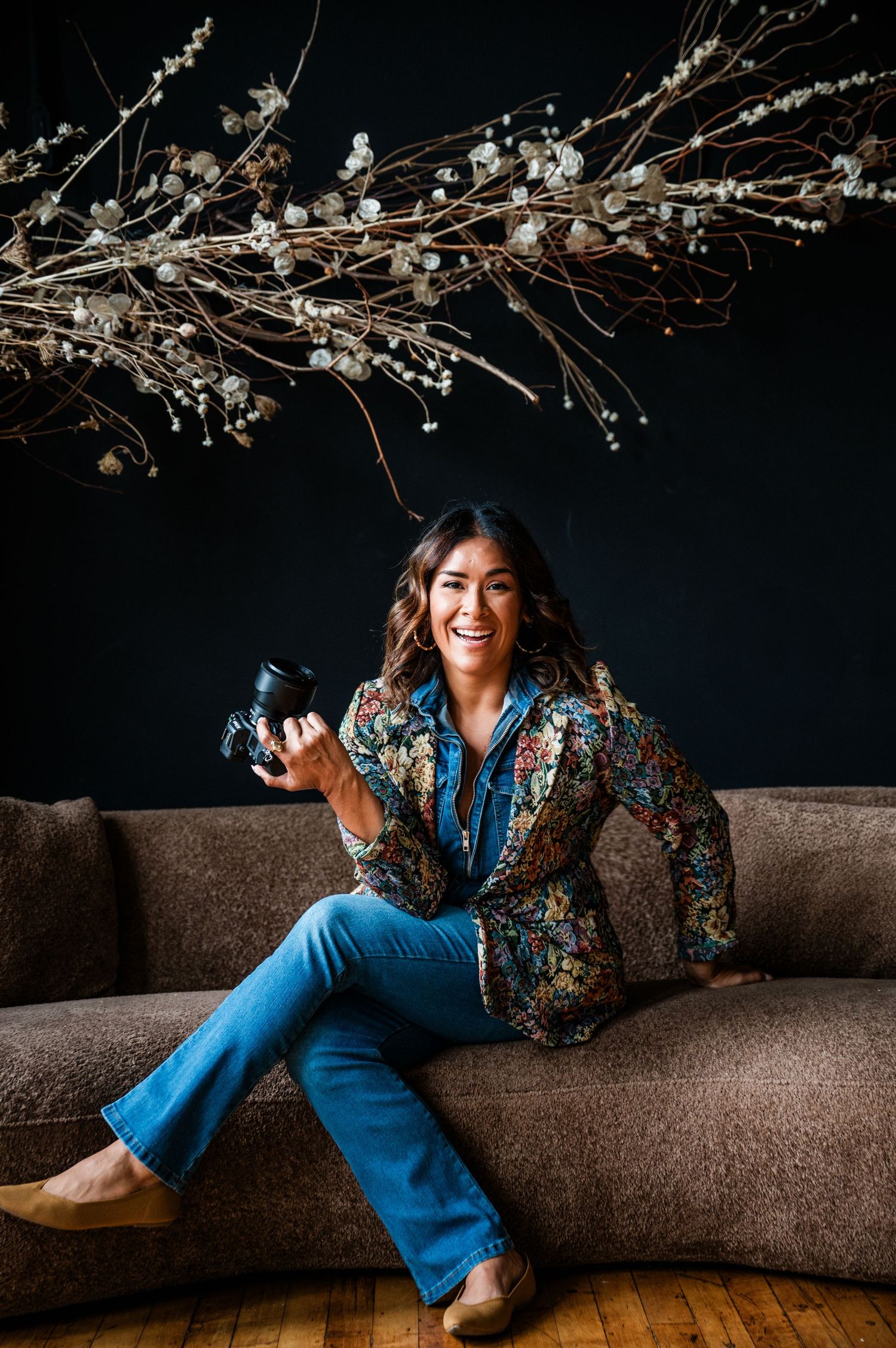Smiling woman in floral blazer and blue jeans sits on sofa holding a Nikon camera, with decorative branches on dark wall in Hartford, CT.