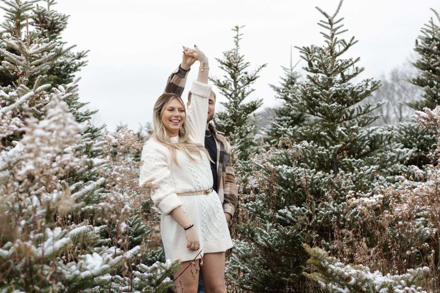 Snowy winter scene at Christmas tree farm with white-clad figures among evergreens.