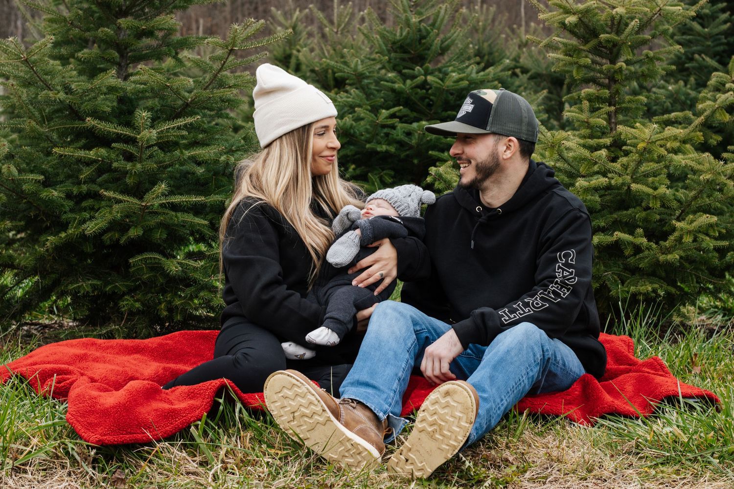 Couple relaxing on red blanket surrounded by Christmas trees during outdoor photoshoot.