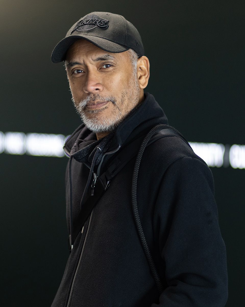 A person wearing a black baseball cap and jacket stands in low lighting against a dark background.