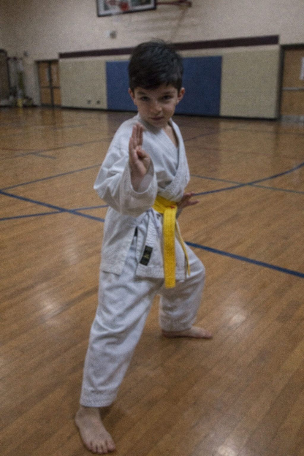 A young martial arts student in white uniform practices kicking techniques in a gymnasium with wooden floors.