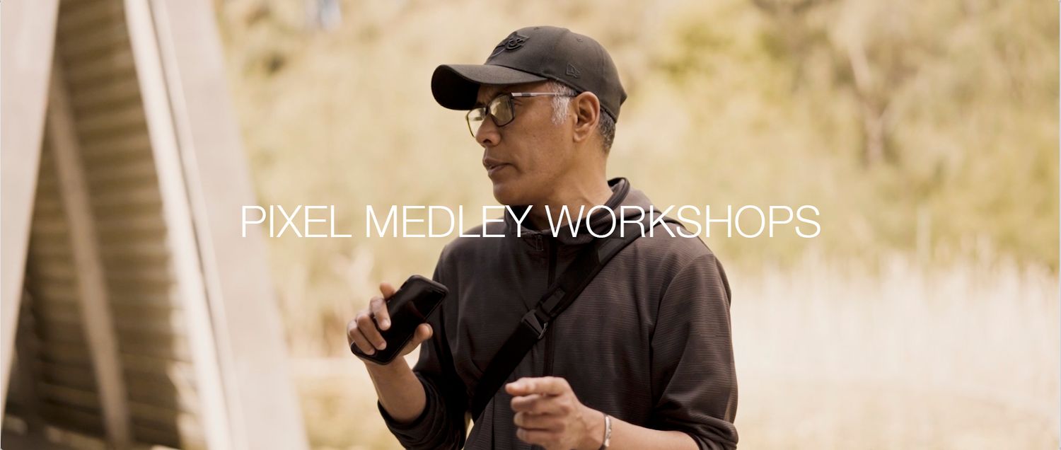 A photographer wearing a black shirt and baseball cap holds a camera while teaching at a Pixel Medley workshop.