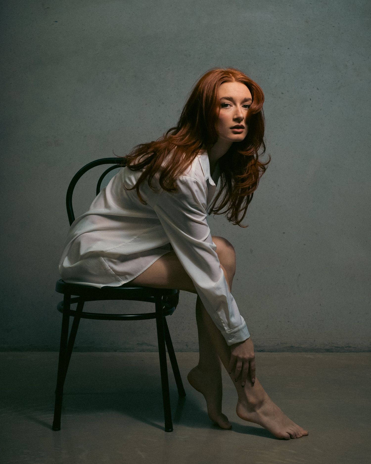 Artistic portrait of a woman with flowing red hair wearing a white shirt while posing on a chair in a minimalist studio setting.