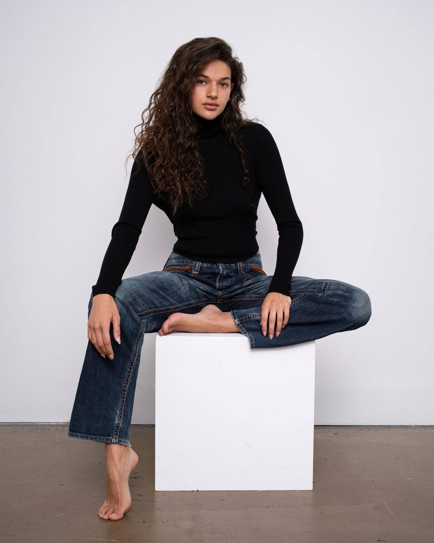 Model in black top and jeans sits casually on white cube against neutral background in studio setting.