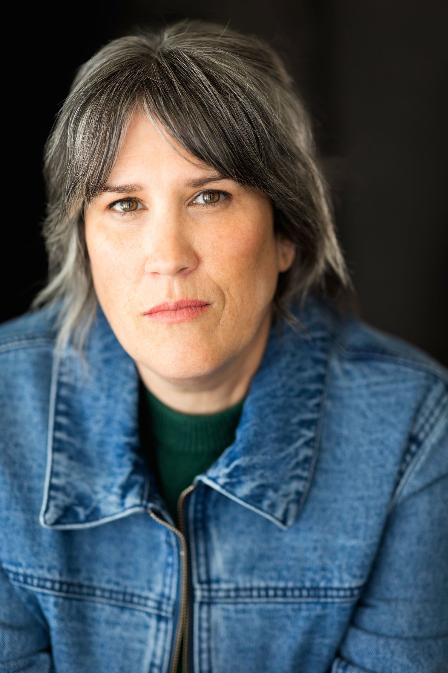Middle-aged woman with gray-streaked hair wearing a denim jacket over green turtleneck, serious expression, portrait photo.