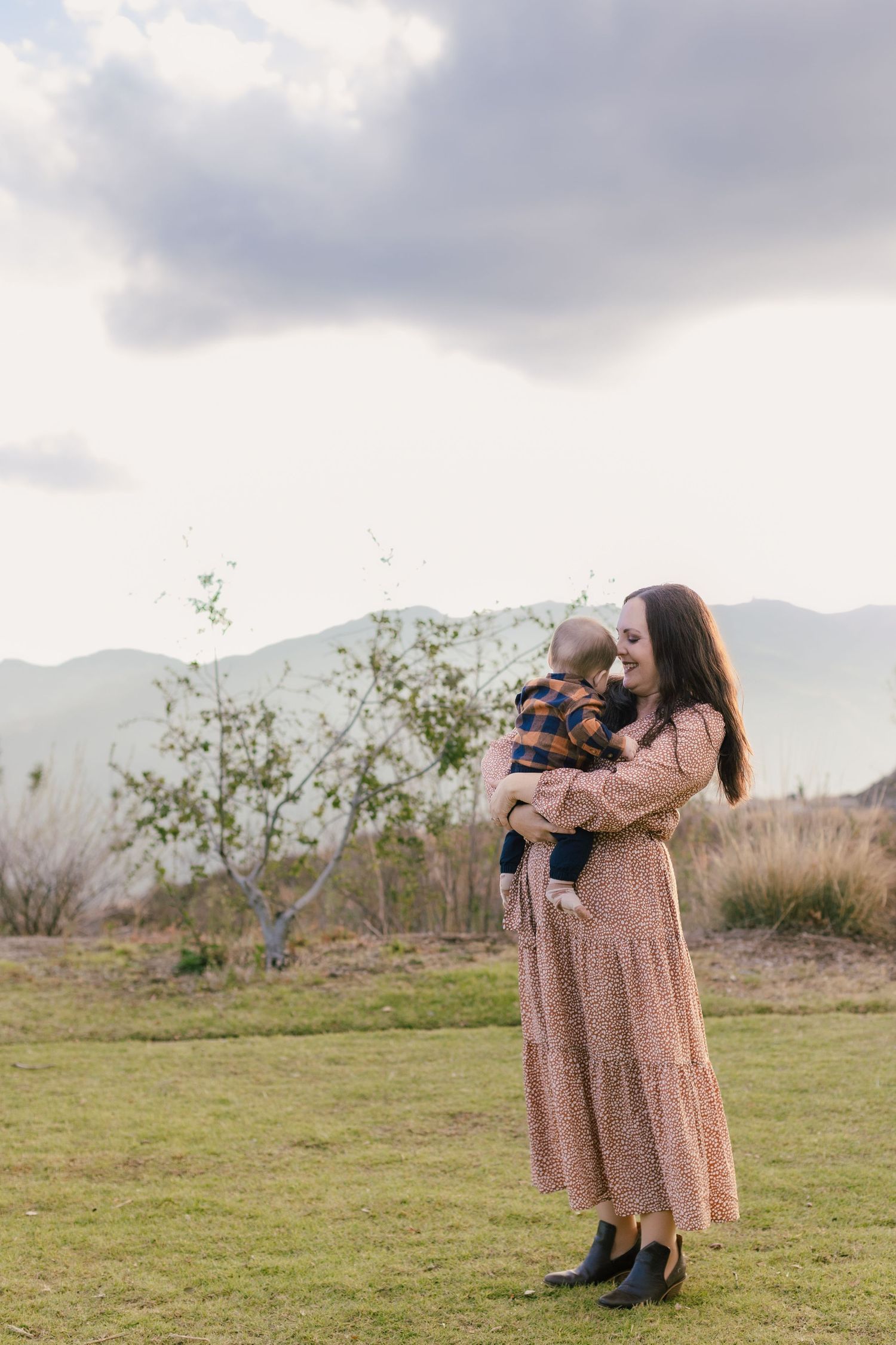Photographer in floral dress capturing moments in an open field with mountains in background.