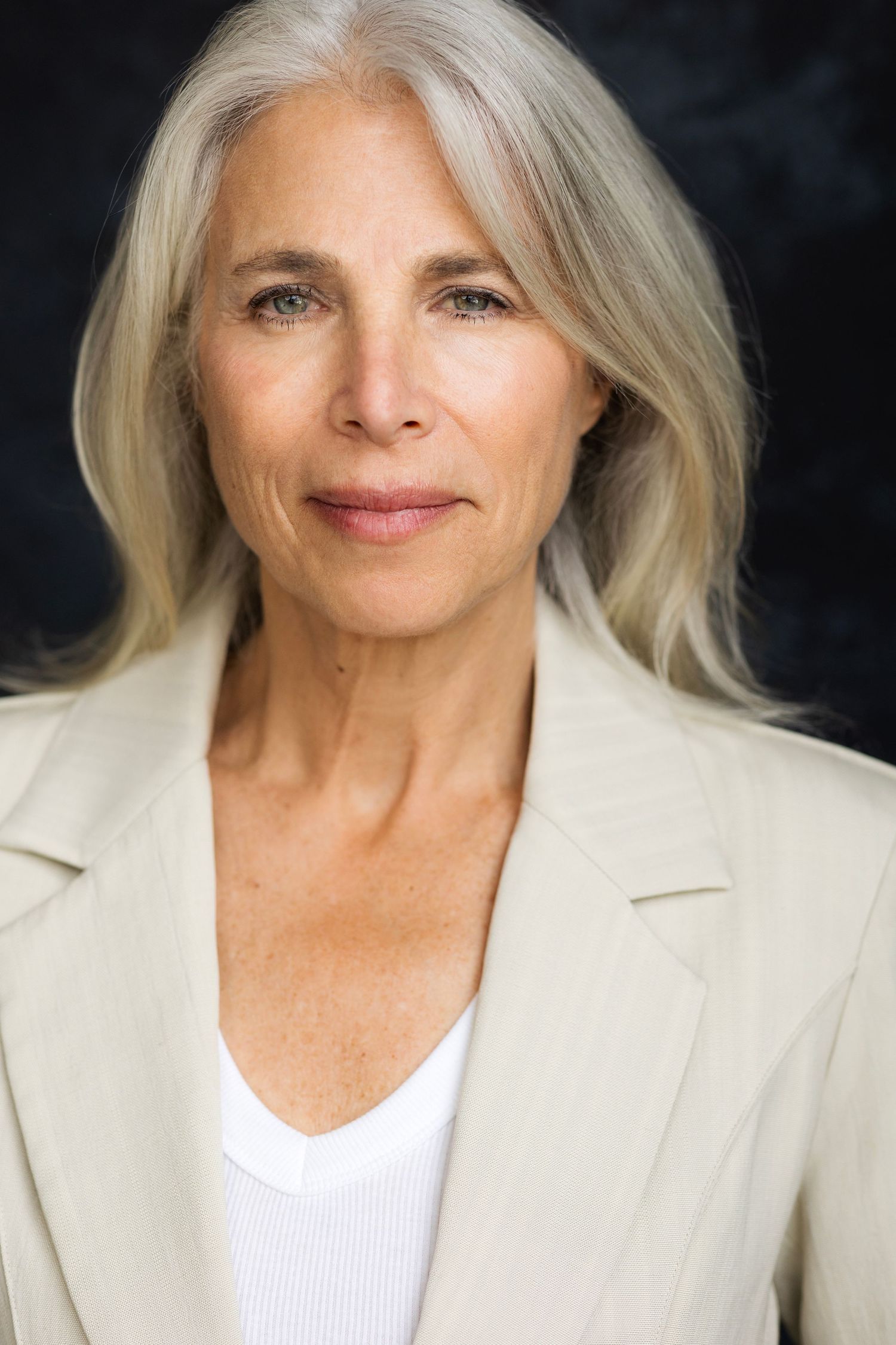 Elegant older woman with silver hair wearing a beige blazer, smiling confidently against a dark background.