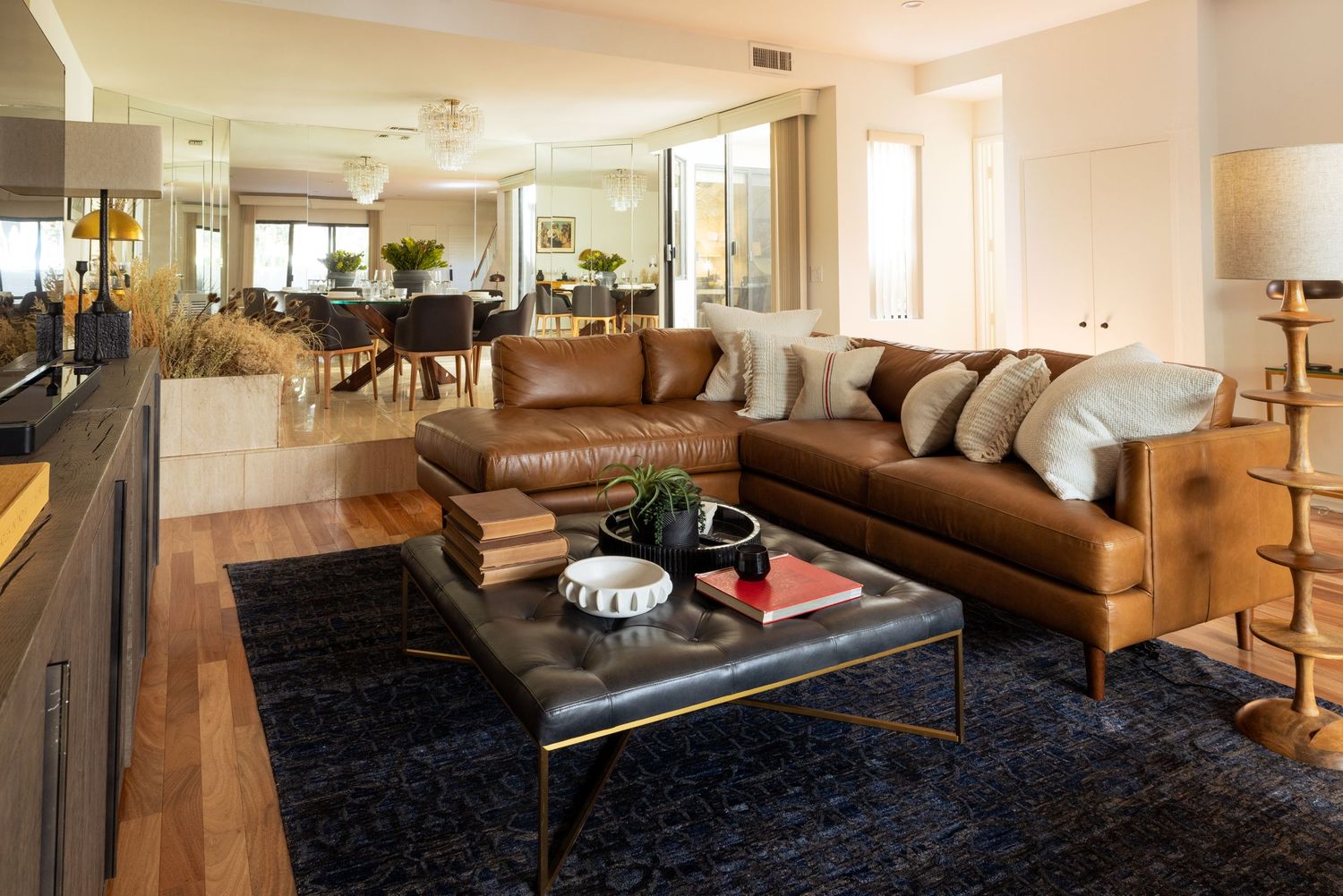 Modern living room with brown leather sectional sofa, dark blue rug, and leather ottoman coffee table with open dining area.
