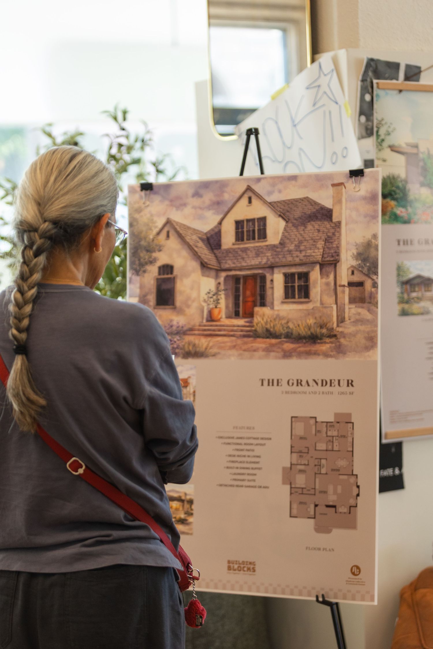 Architectural presentation boards showing house renderings and floor plans displayed on easels in a bright office setting.