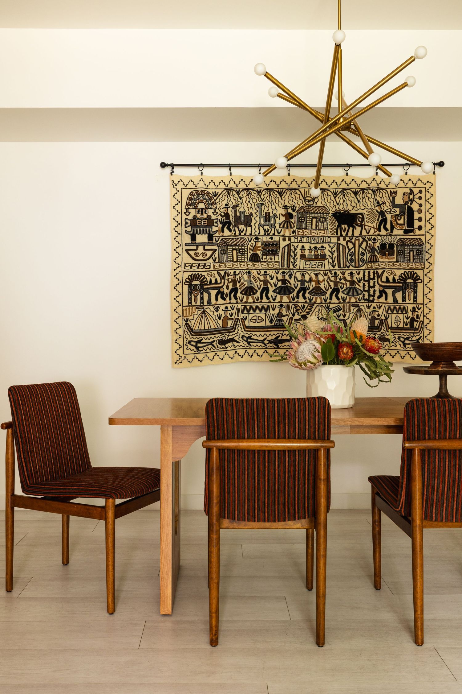Mid-century dining room with walnut table, striped brown chairs, sputnik chandelier, and folk art tapestry on white wall.
