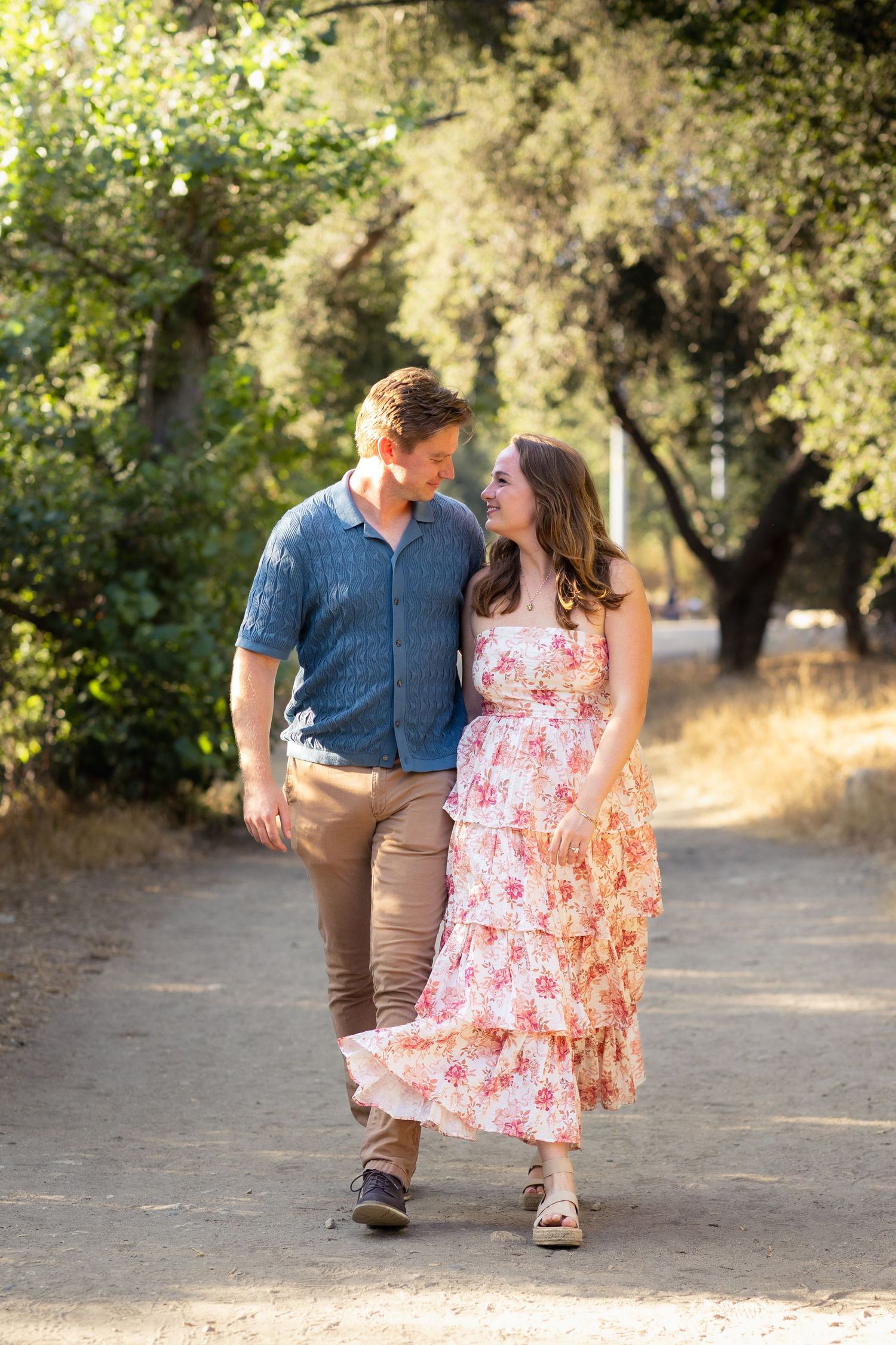A couple walking together on a path, with her wearing a flowing floral dress in summer sunlight.